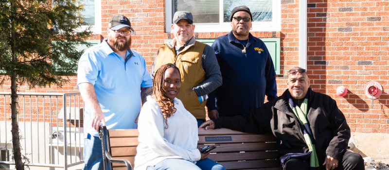 Three men standing and a women and man sitting on a bench.